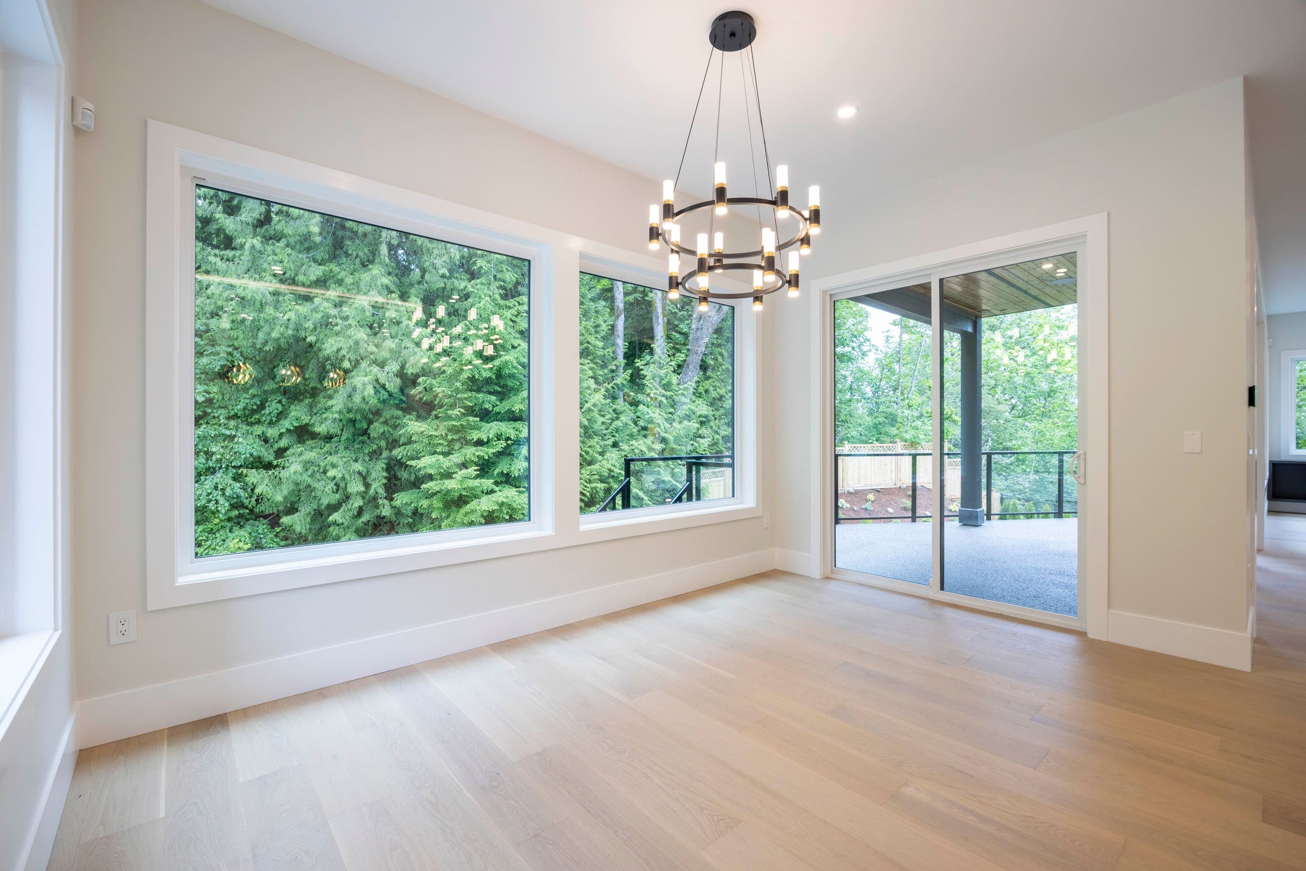 Dining area beneath a ring chandelier, forest through the corner glazing — The Monashee House